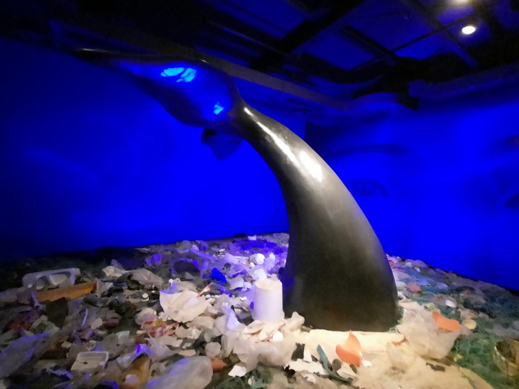 Nachgebildete Schwanzflosse eines Wals, die aus einem Meer voller Kunststoff herausragt.
