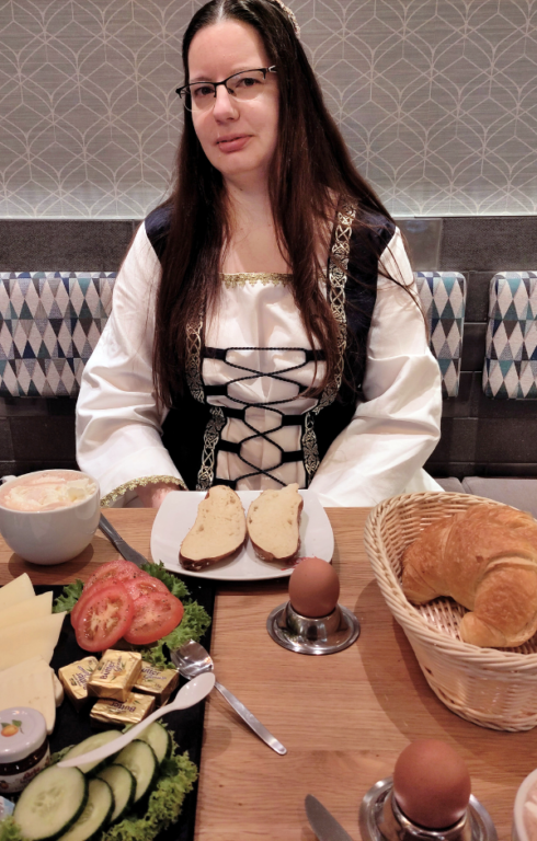 Eine Frau (Rosi) sitzt, in mittelalterlicher Gewandung, beim Bäcker und frühstückt.