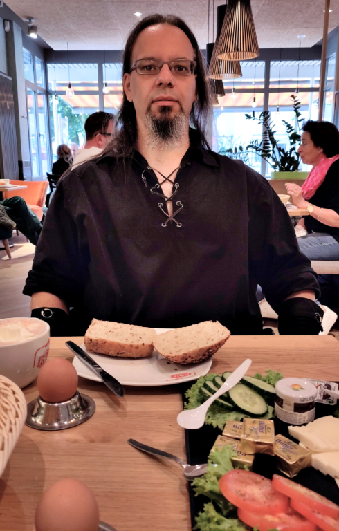 Ein Mann (Ron) sitzt, in mittelalterlicher Gewandung, beim Bäcker und frühstückt.
