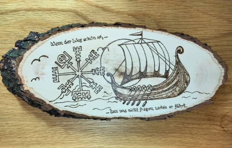 Eine Holzscheibe mit einem Drachenboot auf dem Wasser. Links daneben ein Wikingerkompass. Außerdem der Spruch: "Wenn der Weg schön ist, lass uns nicht fragen wohin er führt."