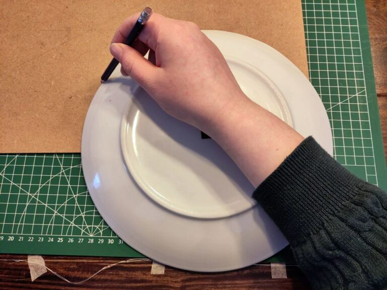 Eine Hand, die den Halbkreis von einem Teller auf eine Hartfaserplatte zeichnet.