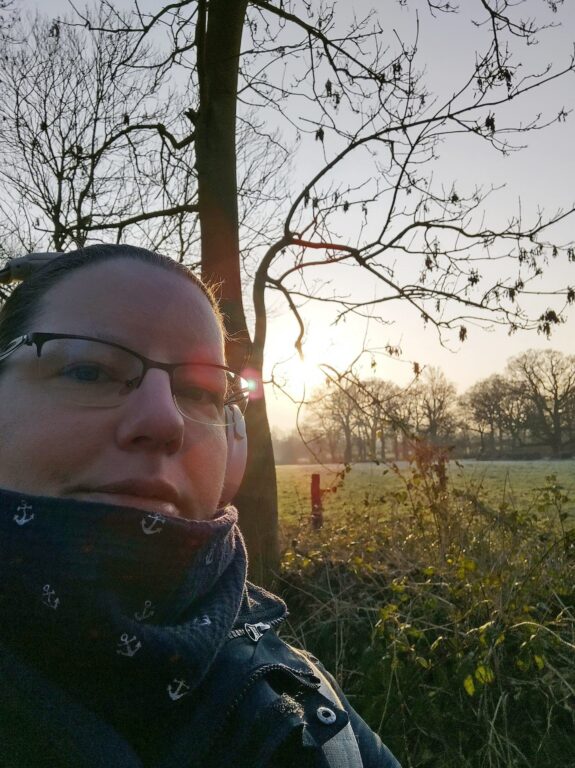Ein Selfie einer Frau um die 40 (Rosi), vor einem Feld, über dem die Sonne aufgeht.