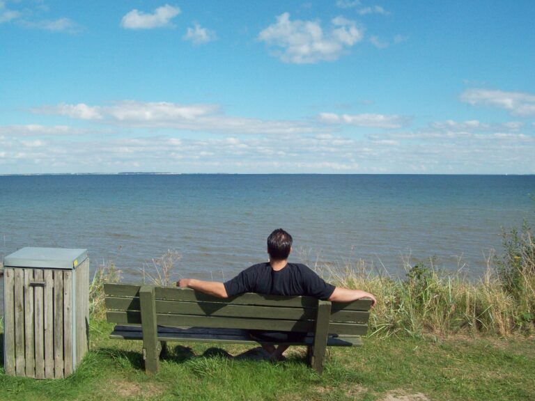 Ein Mann (Ron) sitzt auf einer Bank, vor ihm nichts als das Meer.